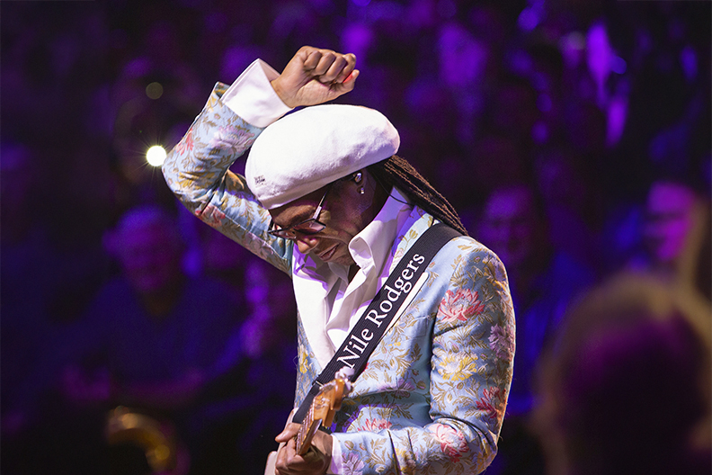 Nile Rogers holding guitar on stage, lifting a fist over his head